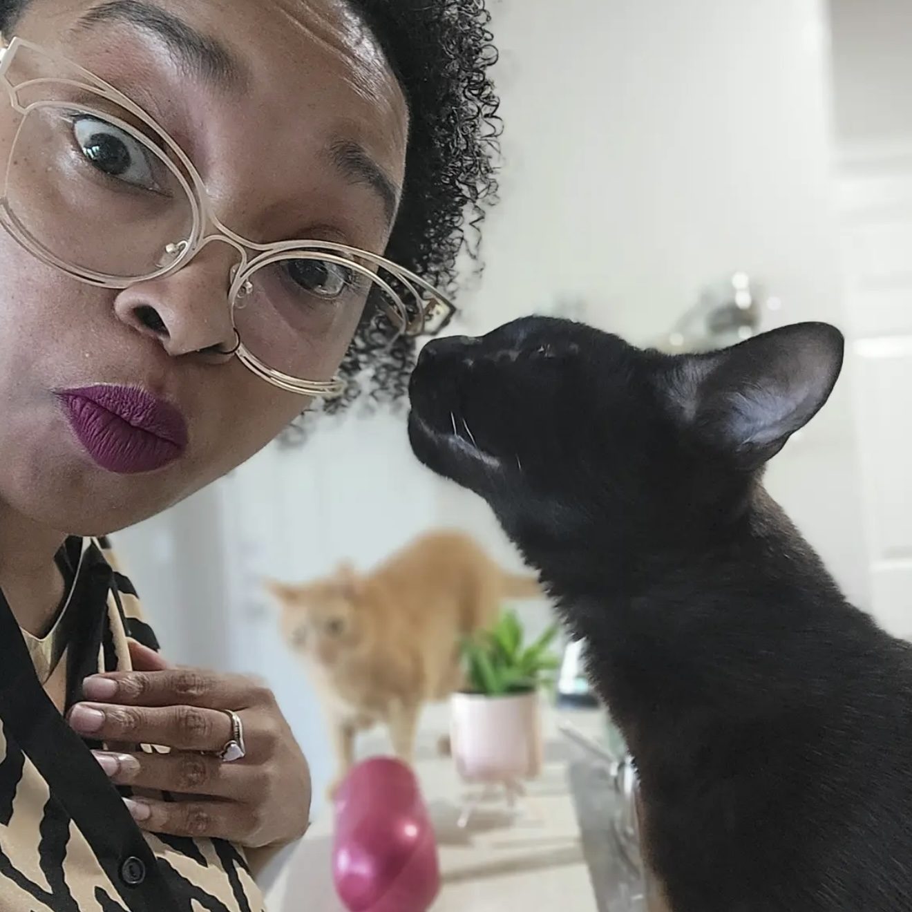 A woman poses playfully with a black cat, while an orange cat is in the background.