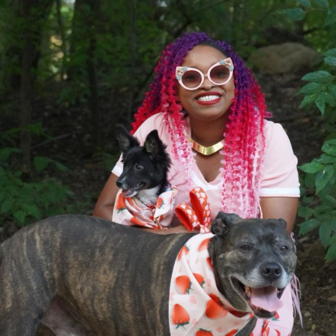 A woman with vibrant hair poses outdoors with two dogs dressed in matching scarves.