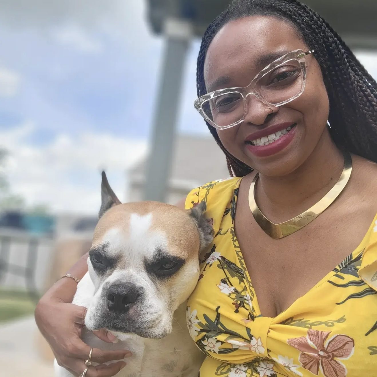 Smiling woman in a yellow floral top holding a dog outdoors.