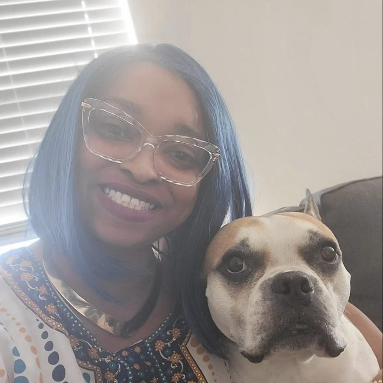 Smiling person with blue hair and glasses, holding a dog with a white and brown coat.